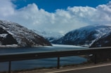A scenic view of a fjord bordered by snow-capped mountains with a body of water beneath a cloudy sky. The foreground features a road with a metal guardrail, suggesting travel through a mountainous landscape.