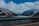 A scenic view of a fjord bordered by snow-capped mountains with a body of water beneath a cloudy sky. The foreground features a road with a metal guardrail, suggesting travel through a mountainous landscape.