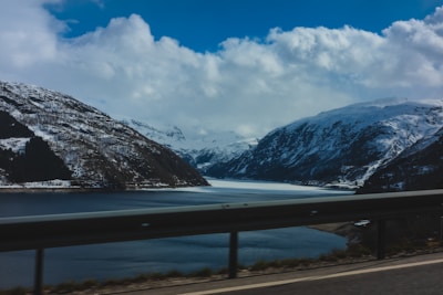 A scenic view of a fjord bordered by snow-capped mountains with a body of water beneath a cloudy sky. The foreground features a road with a metal guardrail, suggesting travel through a mountainous landscape.