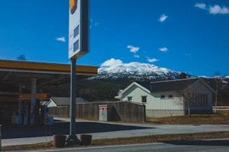 A gas station with a Shell logo is situated on the left, featuring fuel pumps and an overhanging canopy. In the background, snow-capped mountains stand under a clear blue sky, while a house with light-colored walls and a dark roof is on the right, bordered by a wooden fence.
