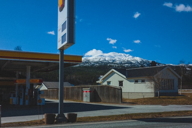 A gas station with a Shell logo is situated on the left, featuring fuel pumps and an overhanging canopy. In the background, snow-capped mountains stand under a clear blue sky, while a house with light-colored walls and a dark roof is on the right, bordered by a wooden fence.