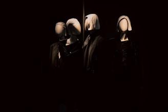 A dimly lit display featuring three mannequins dressed in dark clothing with smooth, featureless faces. The mannequins are arranged side by side, creating a dramatic and mysterious atmosphere in the surrounding darkness.