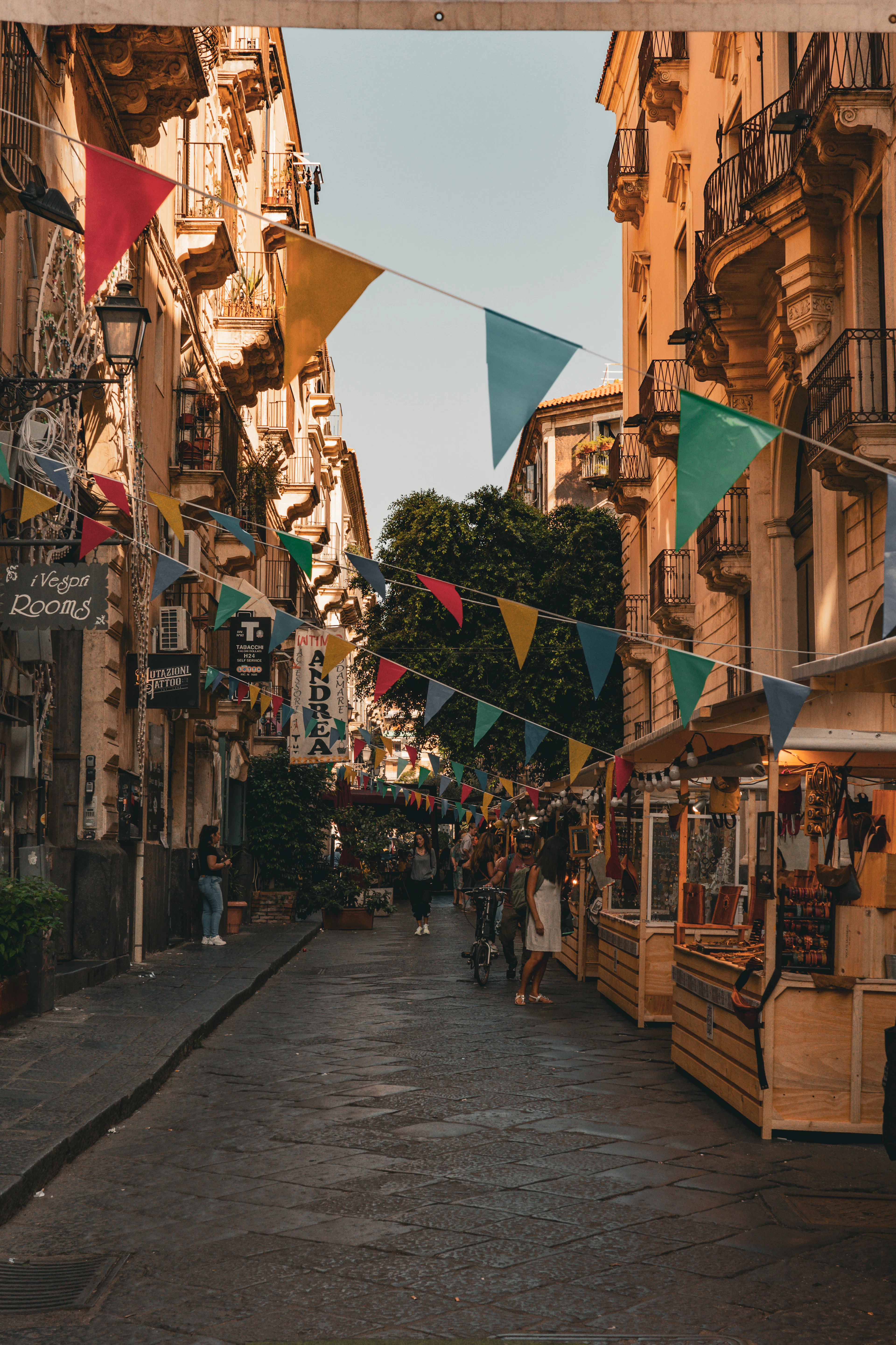 Colorful bunting flags hang above a narrow Catania lane lined with market stalls at dusk.