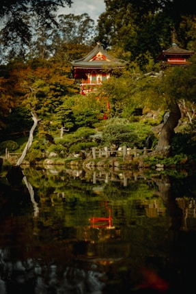 Beautiful oriental-style pagoda structure surrounded by lush greenery and trees with their reflection in the calm water below. The scene conveys serenity and natural beauty, with a well-balanced combination of architecture and nature.