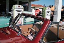 A vintage red convertible is parked in front of retro-style gasoline pumps. The pumps have labels that read 'Flying Gasoline.' A green classic car is visible behind the pumps, and there's a large Coca-Cola sign in the background. The scene includes several people, some of whom are standing near the pumps and others are seated.