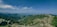 A panoramic view of lush green mountains under a clear blue sky. Dense forests cover the rolling hills, and rocky outcrops are scattered throughout the landscape.