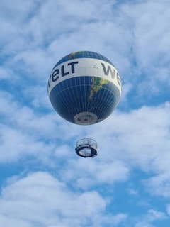 A large, spherical hot air balloon with a depiction of the Earth and the word 'WELT' printed on its sides is floating in a bright blue sky with scattered white clouds. The basket beneath the balloon is suspended by ropes.