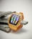 A smartwatch with a vibrant orange strap and a yellow, gem-embedded frame displaying colorful app icons on its screen. It rests on a stack of flat, gray stones against a neutral background.