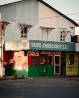 A street corner features a small taxi service building with a worn, corrugated metal upper section and signage for 'TAXIS CHIRICANOS S.A.'. The building's exterior is painted in green and red, with a barber shop to the right, indicated by a yellow sign. There are various windows and a door with visible bars and an urban, slightly rundown appearance.