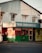 A street corner features a small taxi service building with a worn, corrugated metal upper section and signage for 'TAXIS CHIRICANOS S.A.'. The building's exterior is painted in green and red, with a barber shop to the right, indicated by a yellow sign. There are various windows and a door with visible bars and an urban, slightly rundown appearance.