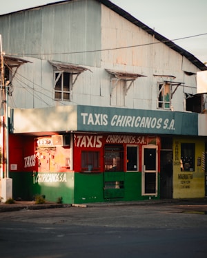 A street corner features a small taxi service building with a worn, corrugated metal upper section and signage for 'TAXIS CHIRICANOS S.A.'. The building's exterior is painted in green and red, with a barber shop to the right, indicated by a yellow sign. There are various windows and a door with visible bars and an urban, slightly rundown appearance.