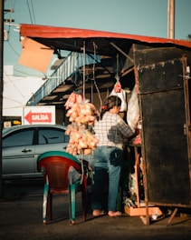 A vendor at an outdoor market stall is tending to her goods. There are bags of oranges hanging from the roof, and a variety of other items are displayed. The stall has a makeshift, rustic setup with a simple shelter. Nearby, a car is parked and a sign reading 'SALIDA' is visible, indicating an exit. The lighting suggests late afternoon with long shadows.