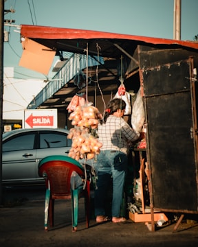 A vendor at an outdoor market stall is tending to her goods. There are bags of oranges hanging from the roof, and a variety of other items are displayed. The stall has a makeshift, rustic setup with a simple shelter. Nearby, a car is parked and a sign reading 'SALIDA' is visible, indicating an exit. The lighting suggests late afternoon with long shadows.