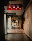A dimly lit, narrow shopping aisle with stores on either side. Most of the store entrances are covered with metal shutters, suggesting the shops are closed. There are various signs hanging from the ceiling advertising different shops and promotions, including a prominent red sign for Premium Outlet displaying discounted prices for various days of the week.