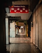 A dimly lit, narrow shopping aisle with stores on either side. Most of the store entrances are covered with metal shutters, suggesting the shops are closed. There are various signs hanging from the ceiling advertising different shops and promotions, including a prominent red sign for Premium Outlet displaying discounted prices for various days of the week.