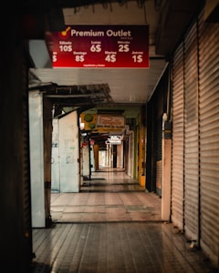 A dimly lit, narrow shopping aisle with stores on either side. Most of the store entrances are covered with metal shutters, suggesting the shops are closed. There are various signs hanging from the ceiling advertising different shops and promotions, including a prominent red sign for Premium Outlet displaying discounted prices for various days of the week.