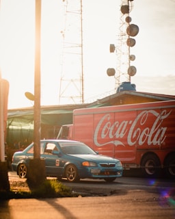 An urban setting with a blue car and a large red delivery truck featuring a Coca-Cola logo. The scene is illuminated by warm, golden sunlight casting long shadows. Behind the vehicles, there are communication towers with multiple dishes and a barbed-wire fence visible on the rooftop.