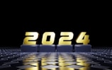 Golden numbers form the year 2024, displayed prominently on a reflective, checkered surface. The background is predominantly black, emphasizing the metallic sheen of the numbers.