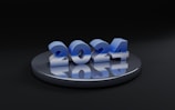 The image features large, metallic blue numbers reading '2024' placed on a round, reflective dark gray platform. The background is predominantly black, enhancing the prominence of the numbers.