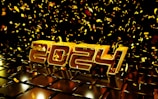 Golden numbers forming '2024' are set against a dark background with a glossy, reflective surface beneath them. Bright golden confetti is scattered throughout the scene, drifting down from above.
