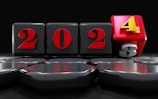 A sequence of four black blocks with the numbers 2023, where the '3' block is rolling over to become '4', indicating the transition to 2024. The numbers are in red with the '4' in yellow on a red block. The background is dark, creating a striking contrast with the glossy blocks.