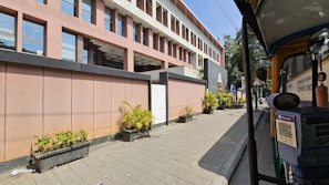A modern building with a geometric design featuring pink and gray tones is adjacent to a sidewalk. There are rectangular sections of windows and potted plants aligned along the pavement. Additionally, an auto rickshaw is parked on the right side of the image.