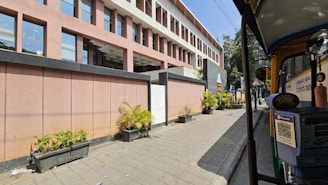 A modern building with a geometric design featuring pink and gray tones is adjacent to a sidewalk. There are rectangular sections of windows and potted plants aligned along the pavement. Additionally, an auto rickshaw is parked on the right side of the image.