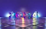 Large 3D numbers '2024' stand on a reflective tiled surface, illuminated by colorful lighting that casts a gradient across the scene. The background features a smooth gradient transitioning from blue to purple, complementing the shiny metallic look of the numbers.