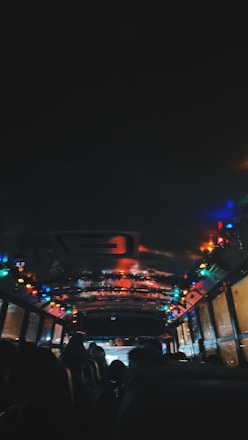 The interior of a dimly lit bus is decorated with colorful string lights along the ceiling. Passengers are seated, and their silhouettes are visible against the brightness of the lights. The atmosphere appears festive, suggesting a holiday or celebration theme.