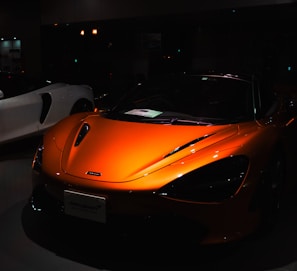A sleek orange sports car is prominently displayed, with its shiny surface reflecting light subtly. The car's design features smooth, aerodynamic curves and distinctive headlights. In the dimly lit background, a white sports car is partially visible.