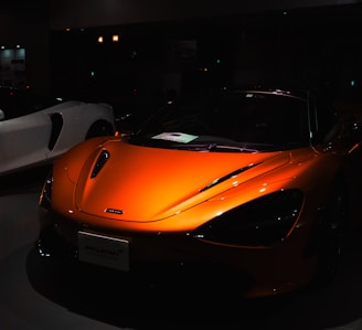 A sleek orange sports car is prominently displayed, with its shiny surface reflecting light subtly. The car's design features smooth, aerodynamic curves and distinctive headlights. In the dimly lit background, a white sports car is partially visible.