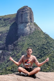 A person sitting cross-legged on a rocky surface, engaging in a meditation pose with hands forming a mudra. The background features a large, impressive rock formation with lush green vegetation covering its slopes under a clear blue sky.