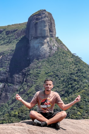 A person sitting cross-legged on a rocky surface, engaging in a meditation pose with hands forming a mudra. The background features a large, impressive rock formation with lush green vegetation covering its slopes under a clear blue sky.