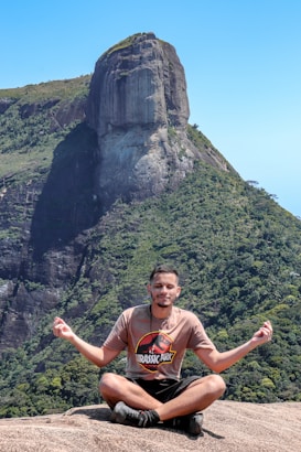 A person sitting cross-legged on a rocky surface, engaging in a meditation pose with hands forming a mudra. The background features a large, impressive rock formation with lush green vegetation covering its slopes under a clear blue sky.