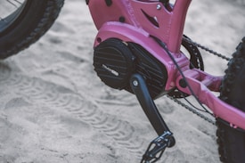 A close-up view of a pink bicycle's drivetrain and pedal on a sandy surface. The bike appears to have mountain bike features, with thick tires and a robust frame. The sand beneath shows tire tracks, indicating movement through the sandy terrain.