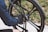 A person in a blue jacket is adjusting the disc brake of a bicycle wheel, focusing on the intricate mechanism. The bicycle has a sleek, modern design with black spokes and a well-maintained tire. The background is slightly blurred, showing a hint of greenery.