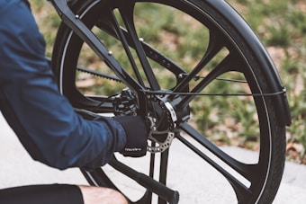 A person in a blue jacket is adjusting the disc brake of a bicycle wheel, focusing on the intricate mechanism. The bicycle has a sleek, modern design with black spokes and a well-maintained tire. The background is slightly blurred, showing a hint of greenery.