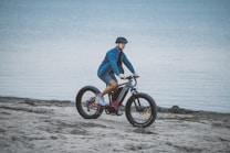 A person is riding a fat-tire bike on a sandy beach near the water. They are wearing a helmet and a blue jacket. The water in the background appears calm and extends to the horizon.