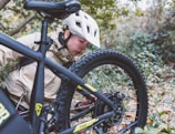 A person wearing a white helmet and beige jacket is closely inspecting the rear tire of a black bicycle. The setting is a natural, forested area with green foliage and fallen leaves on the ground. The focus is on the bicycle's mechanics and the person's attentive posture.