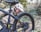 A person wearing a white helmet and beige jacket is closely inspecting the rear tire of a black bicycle. The setting is a natural, forested area with green foliage and fallen leaves on the ground. The focus is on the bicycle's mechanics and the person's attentive posture.