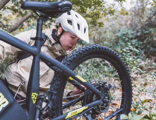 A person wearing a white helmet and beige jacket is closely inspecting the rear tire of a black bicycle. The setting is a natural, forested area with green foliage and fallen leaves on the ground. The focus is on the bicycle's mechanics and the person's attentive posture.