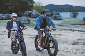 Two men are riding electric bikes on a sandy path near a body of water. Both are wearing helmets, with one in a white helmet and leather jacket, and the other in a black helmet and blue shirt. The background features large rocks, greenery, and mountains in the distance.