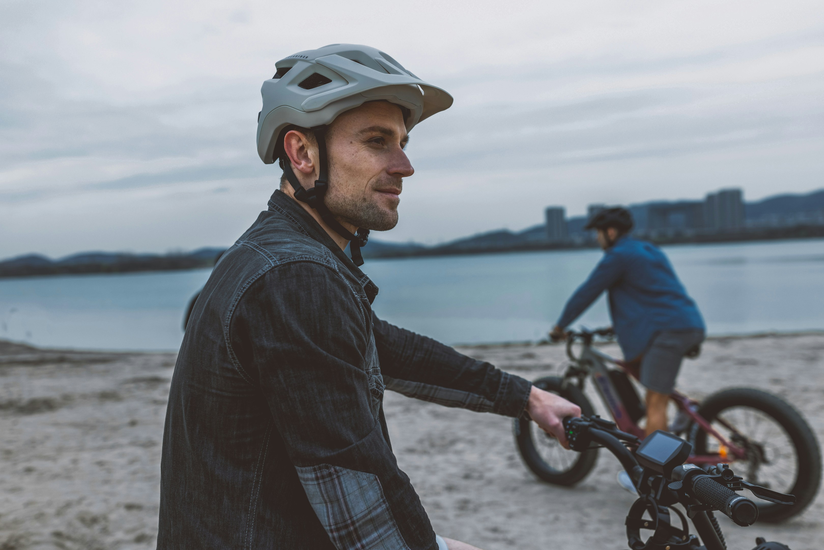 A man wearing a Decathlon helmet sits on an ebike on the beach, looking at his companion