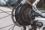A detailed close-up of a bicycle rear wheel, focusing on the gear cassette, derailleur, and disk brake. The metallic components are intricately arranged, showcasing the mechanical design and branding of Shimano and Tourney. Spokes radiate outward from the hub, surrounded by a white bicycle frame in the background.