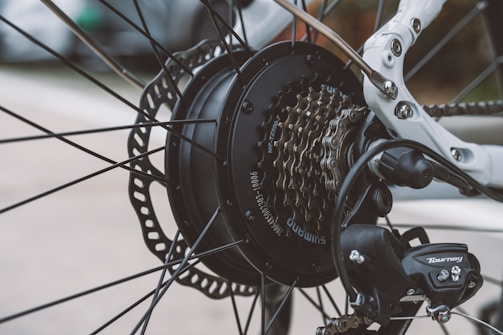 A detailed close-up of a bicycle rear wheel, focusing on the gear cassette, derailleur, and disk brake. The metallic components are intricately arranged, showcasing the mechanical design and branding of Shimano and Tourney. Spokes radiate outward from the hub, surrounded by a white bicycle frame in the background.