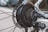 A detailed close-up of a bicycle rear wheel, focusing on the gear cassette, derailleur, and disk brake. The metallic components are intricately arranged, showcasing the mechanical design and branding of Shimano and Tourney. Spokes radiate outward from the hub, surrounded by a white bicycle frame in the background.