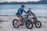 Two people riding electric bikes along a sandy beach with a lake in the background. The riders are wearing casual clothing and helmets, suggesting a leisurely outing. The surrounding environment includes calm water and distant mountains under an overcast sky.