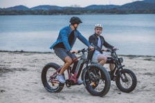 Two people riding electric bikes along a sandy beach with a lake in the background. The riders are wearing casual clothing and helmets, suggesting a leisurely outing. The surrounding environment includes calm water and distant mountains under an overcast sky.