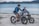 Two people riding electric bikes along a sandy beach with a lake in the background. The riders are wearing casual clothing and helmets, suggesting a leisurely outing. The surrounding environment includes calm water and distant mountains under an overcast sky.