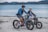 Two people riding electric bikes along a sandy beach with a lake in the background. The riders are wearing casual clothing and helmets, suggesting a leisurely outing. The surrounding environment includes calm water and distant mountains under an overcast sky.